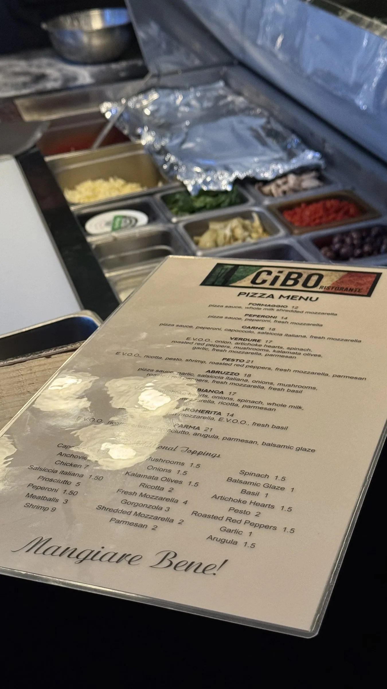 The pizza menu propped on the prep line, with pans of toppings behind it.