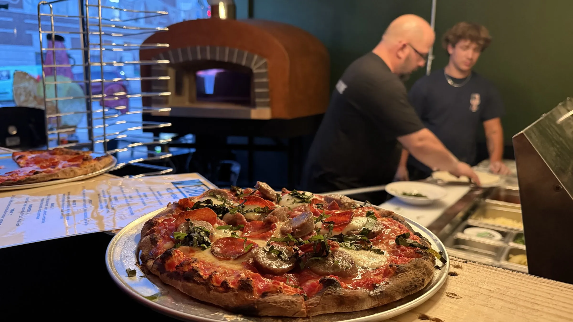 A wood-fired pizza on a tray at the counter, with Wes and the brick oven behind.