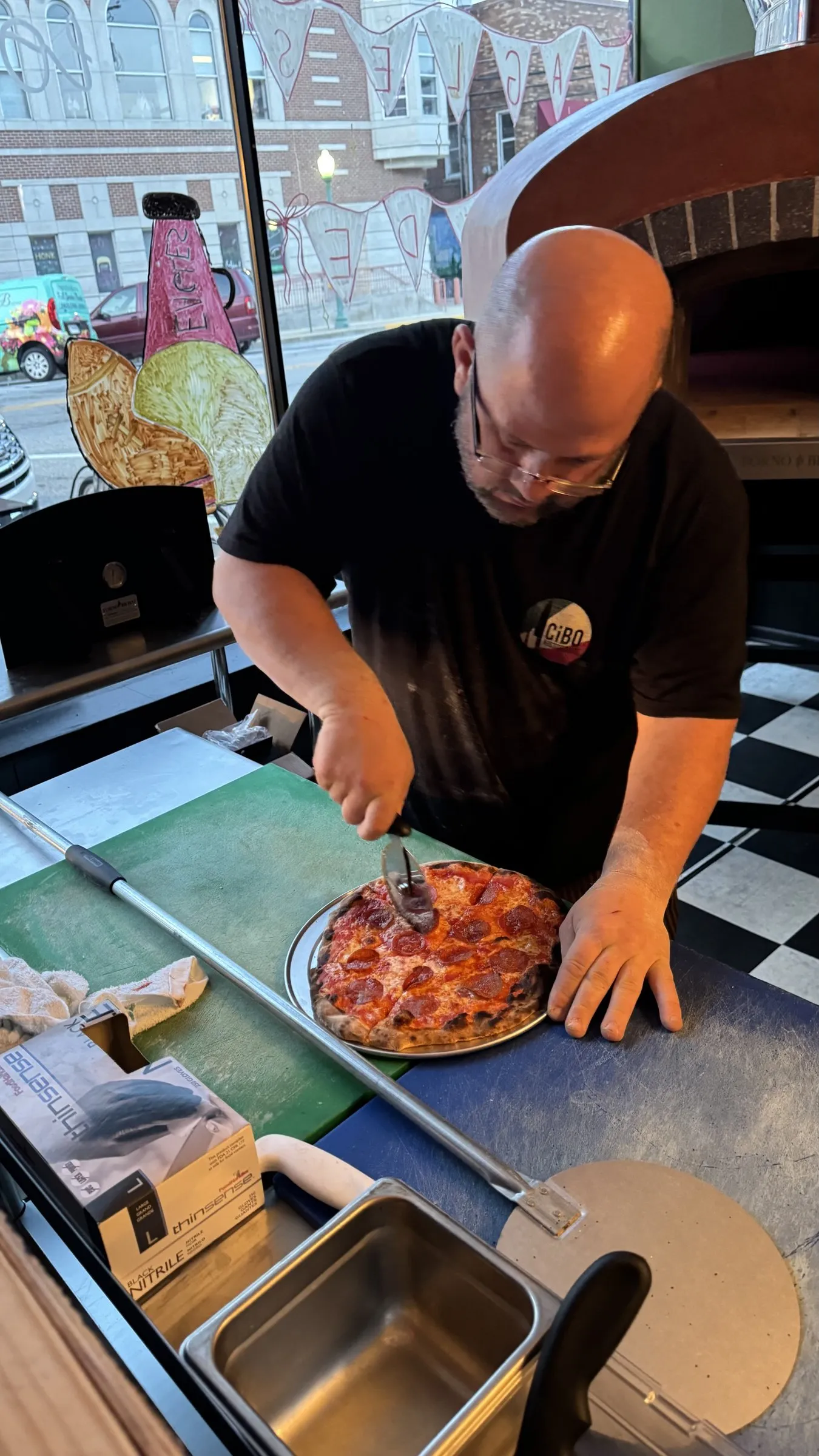 Wes cutting a pepperoni pizza at the counter, the oven and the front window behind him.